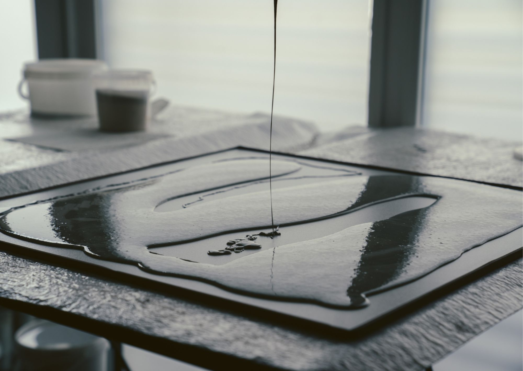 Gros plan d'un liquide versé sur une surface plane et réfléchissante, créant ainsi des motifs abstraits. Deux récipients sont flous à l'arrière-plan et la scène est doucement éclairée par la lumière du jour à travers les fenêtres.