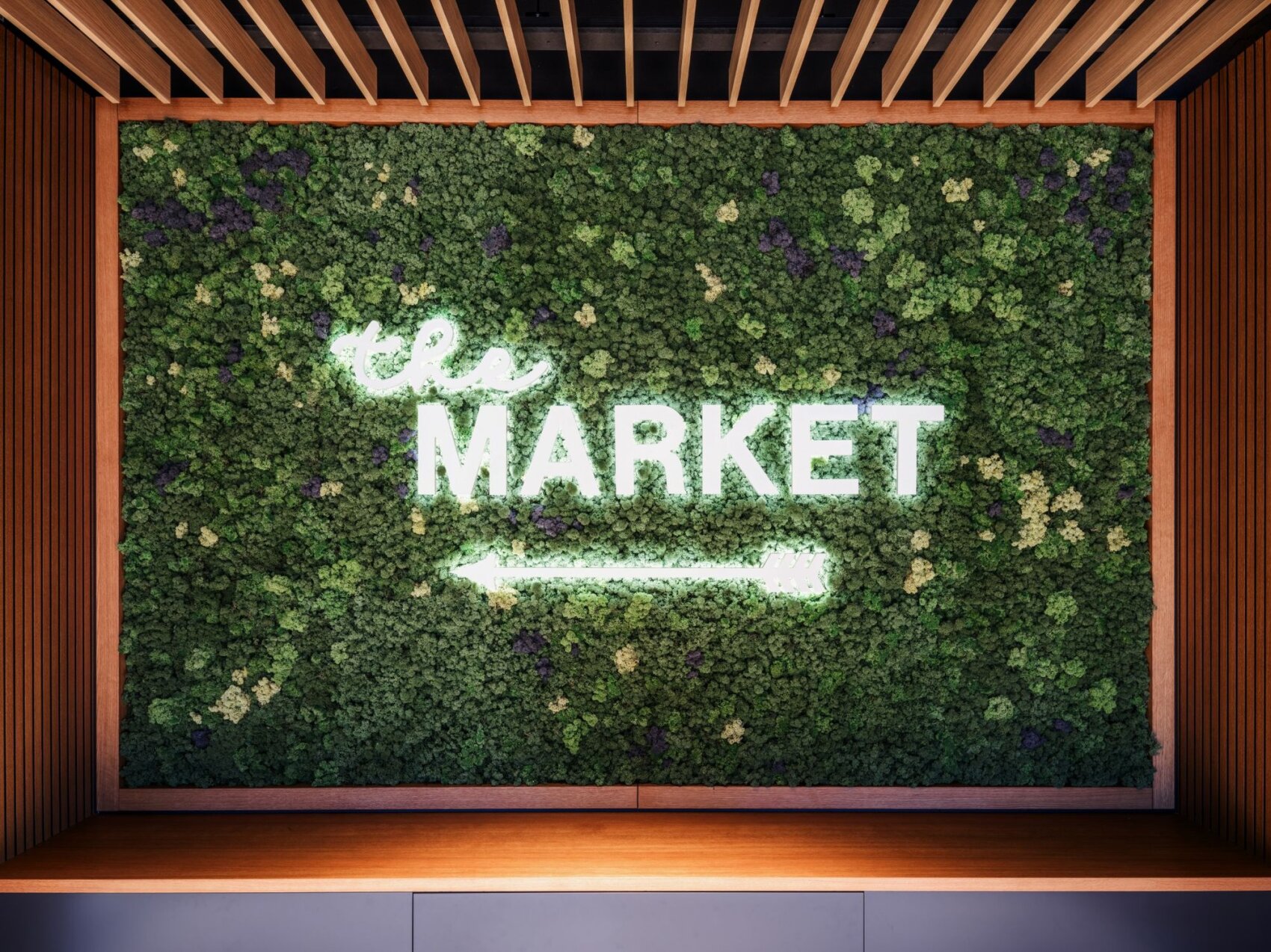 Un mur végétal vert avec une enseigne lumineuse blanche portant l'inscription MARKET et une flèche vers la gauche, encadré de lattes de bois.