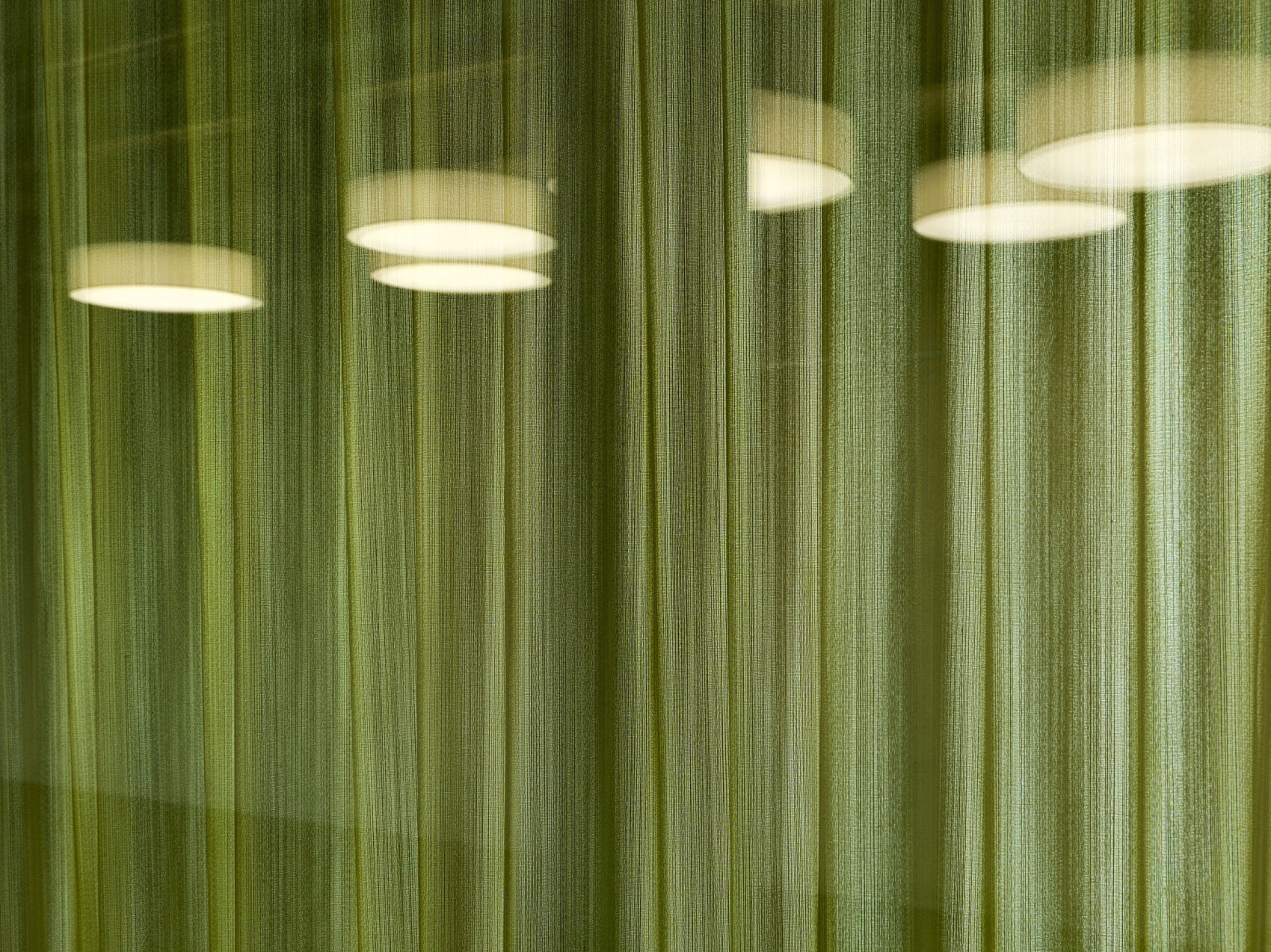 Quatre plafonniers ronds sont visibles à travers un rideau vert semi-transparent, créant un effet doux et diffus avec des lignes verticales provenant de la texture du rideau.