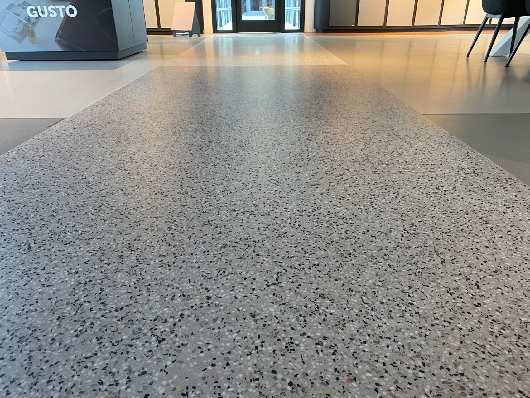 Gros plan sur un sol en terrazzo poli et moucheté dans un bâtiment moderne, avec des tables, des chaises et une grande fenêtre en arrière-plan. La scène est bien éclairée par la lumière naturelle.