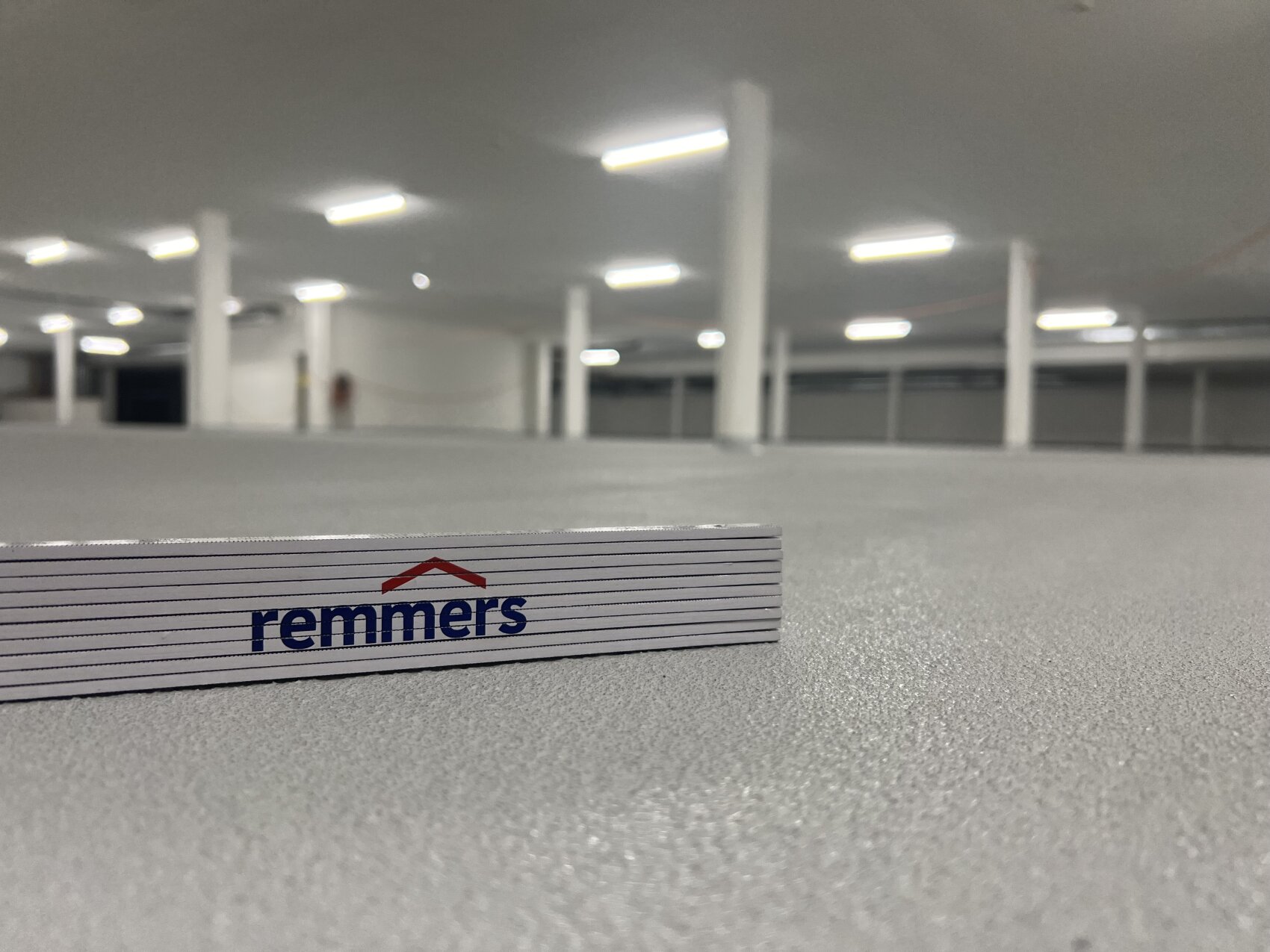 Gros plan d'une règle pliée portant le logo Remmers sur un sol gris texturé dans un intérieur vide, spacieux et bien éclairé, avec des colonnes et des plafonniers blancs.