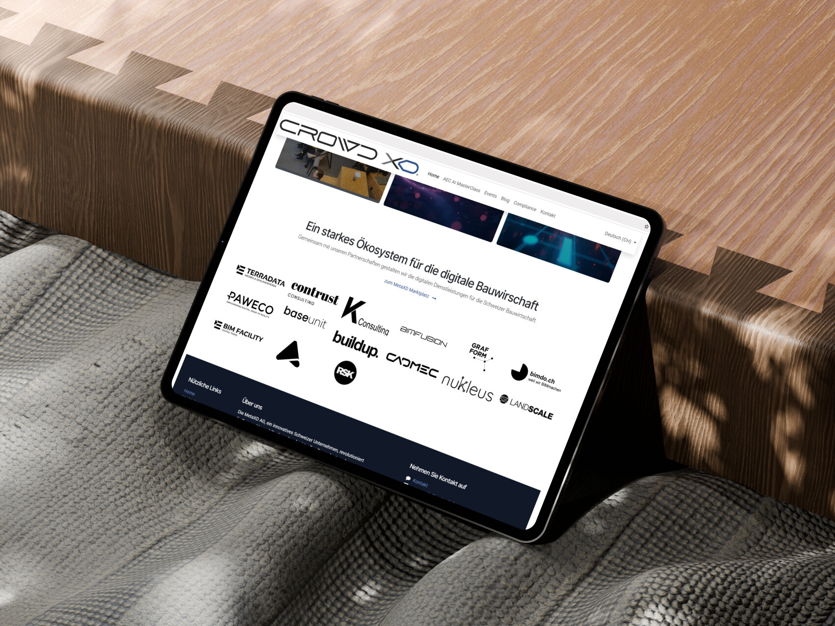 Ein Tablet, auf dem eine Website mit Logos verschiedener Unternehmen angezeigt wird, liegt auf einer grauen, strukturierten Decke und einem Holztisch mit Zickzack-Kantenmuster.