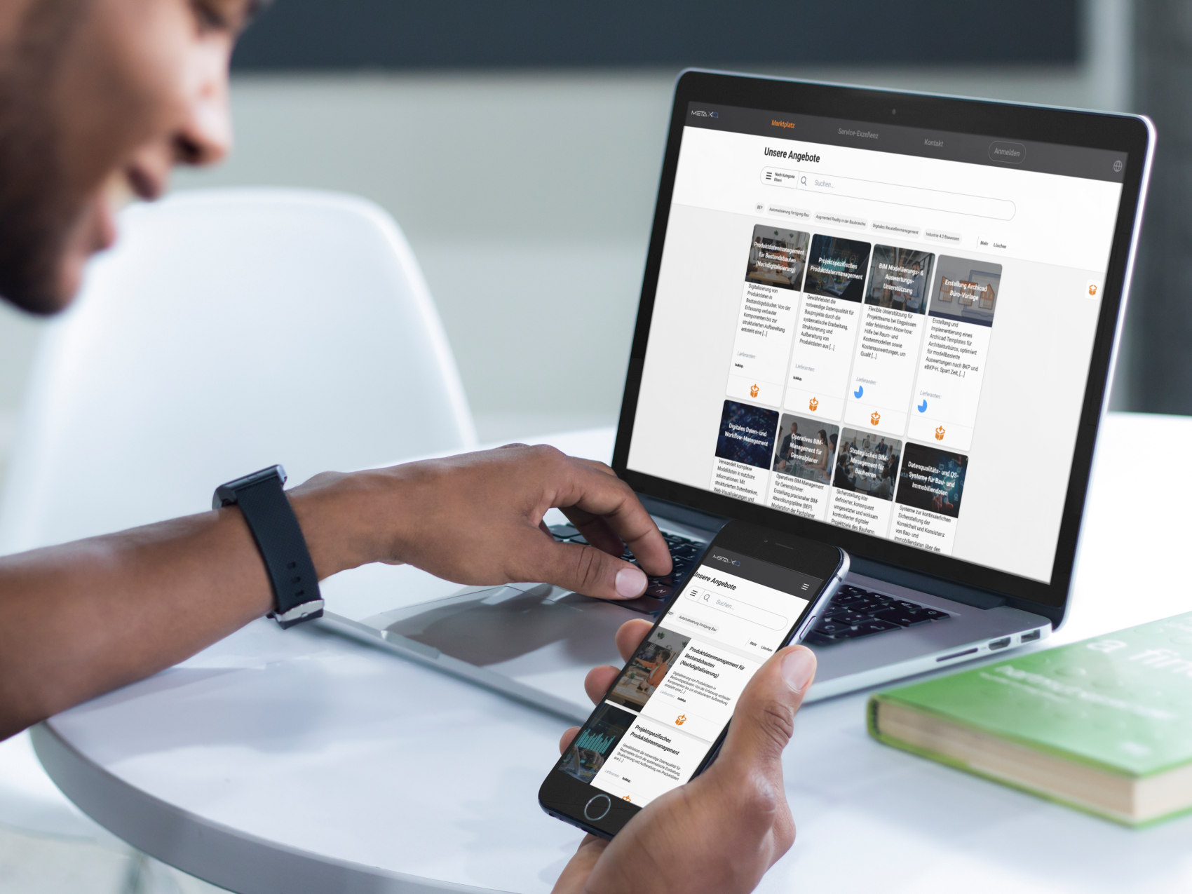 Eine Person sitzt an einem Tisch und benutzt ein Smartphone und einen Laptop, die beide dieselbe Website mit einem Gitter aus Bildern und Text anzeigen. Ein geschlossenes grünes Buch und ein weißer Stuhl stehen ebenfalls auf dem Tisch.