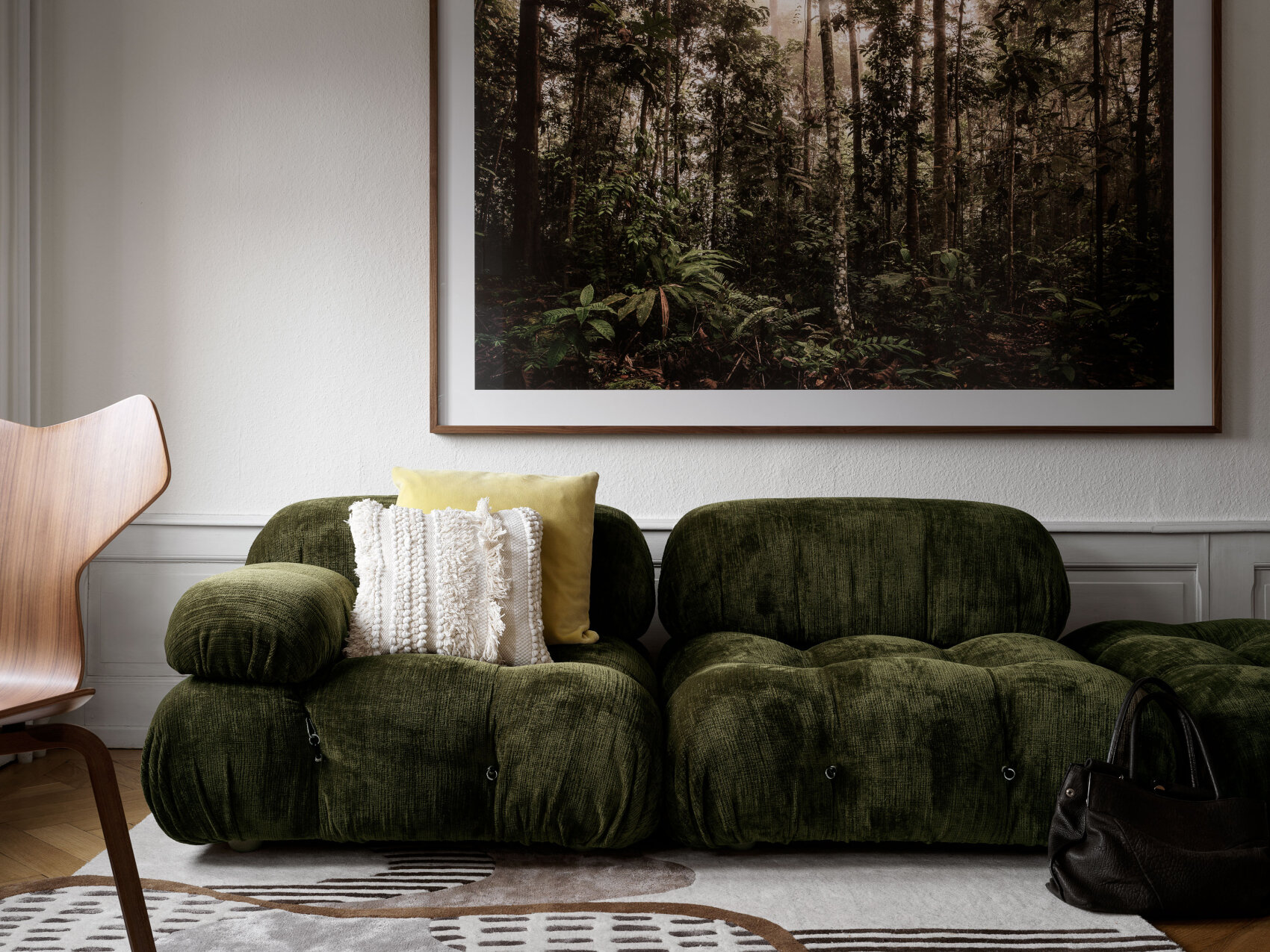 Un salon moderne avec un canapé en velours vert, des coussins décoratifs, un sac à main noir posé sur le sol, une chaise en bois et une grande photographie encadrée d'une forêt luxuriante accrochée au mur au-dessus du canapé.