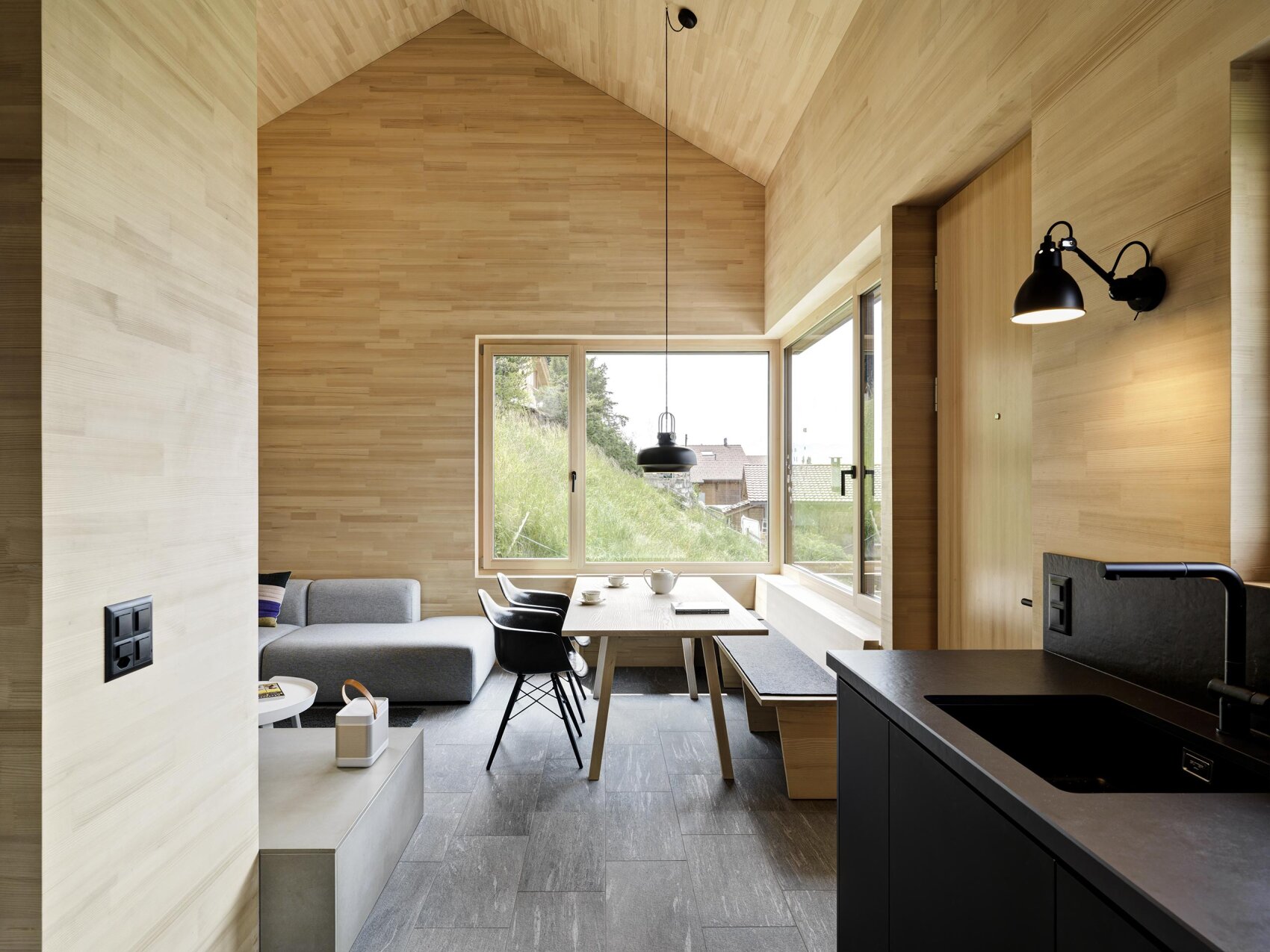 Intérieur minimaliste en bois comprenant un canapé gris moderne, une table à manger avec des chaises noires et un banc, de grandes fenêtres avec vue sur l'extérieur, et une cuisine noire épurée sous une douce lumière naturelle.