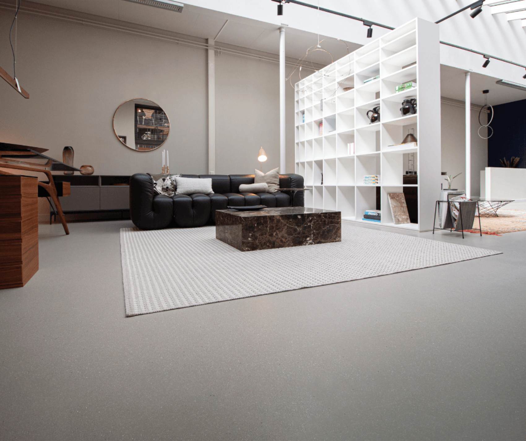 Un salon moderne avec un canapé noir, une table basse en marbre, un tapis blanc et des étagères blanches ouvertes qui divisent la pièce. La pièce est meublée de manière minimaliste et possède de hauts plafonds avec des lucarnes.